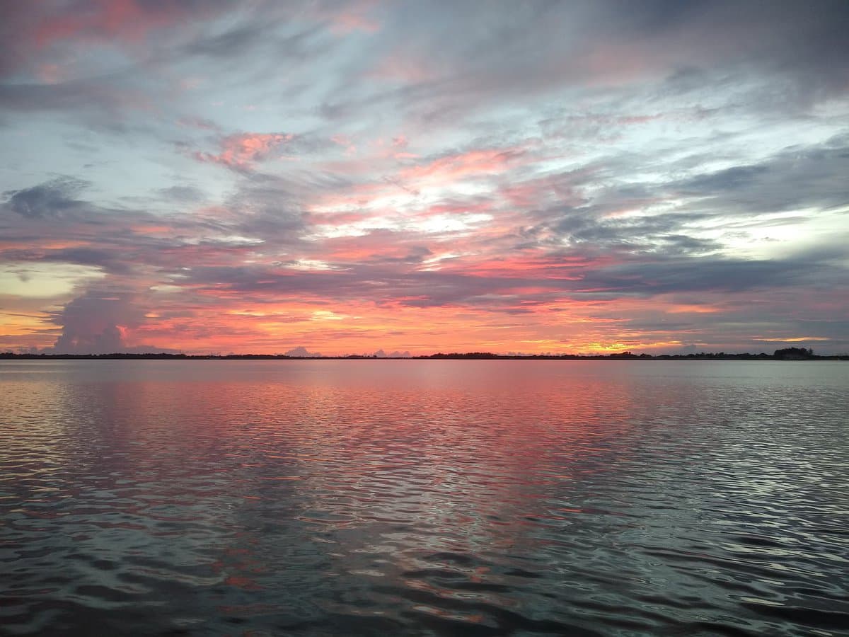 A scenic photograph of a sunset over a calm body of water, with the sky filled with pink, orange, and purple clouds reflecting on the surface. A dark, low-lying shoreline is visible on the horizon.