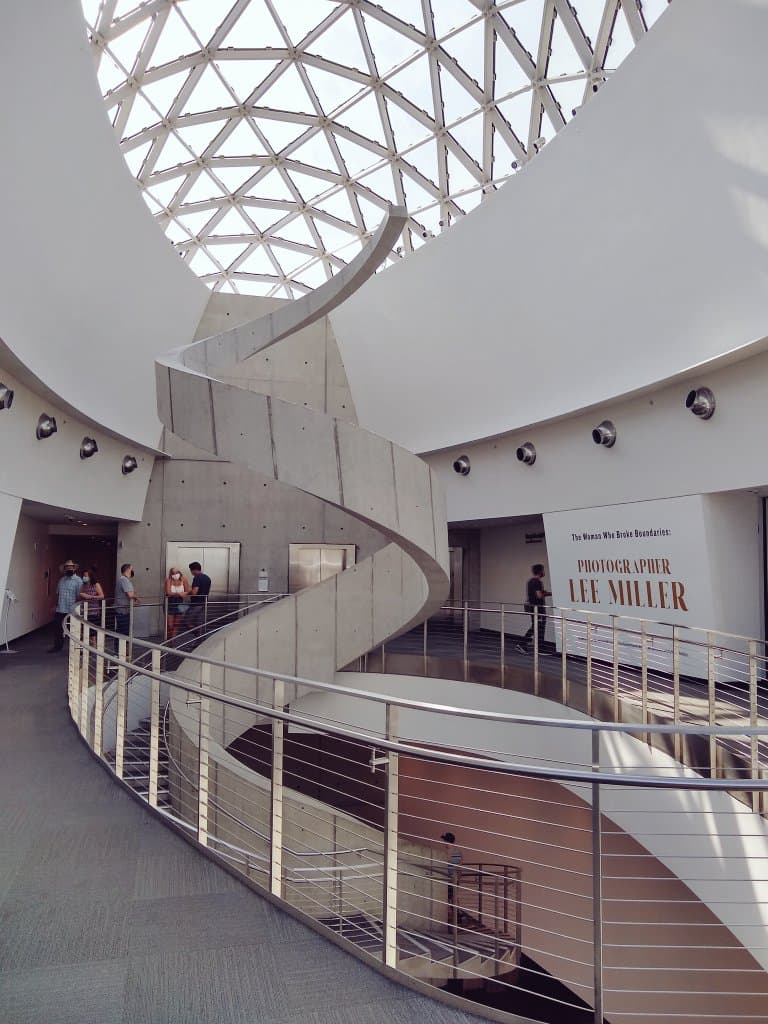 A wide-angle photograph of the interior of a modern museum, featuring a large, white spiral staircase and a glass dome ceiling. On the right wall, a large exhibition sign is visible. Several people are standing on the upper and lower levels of the building.