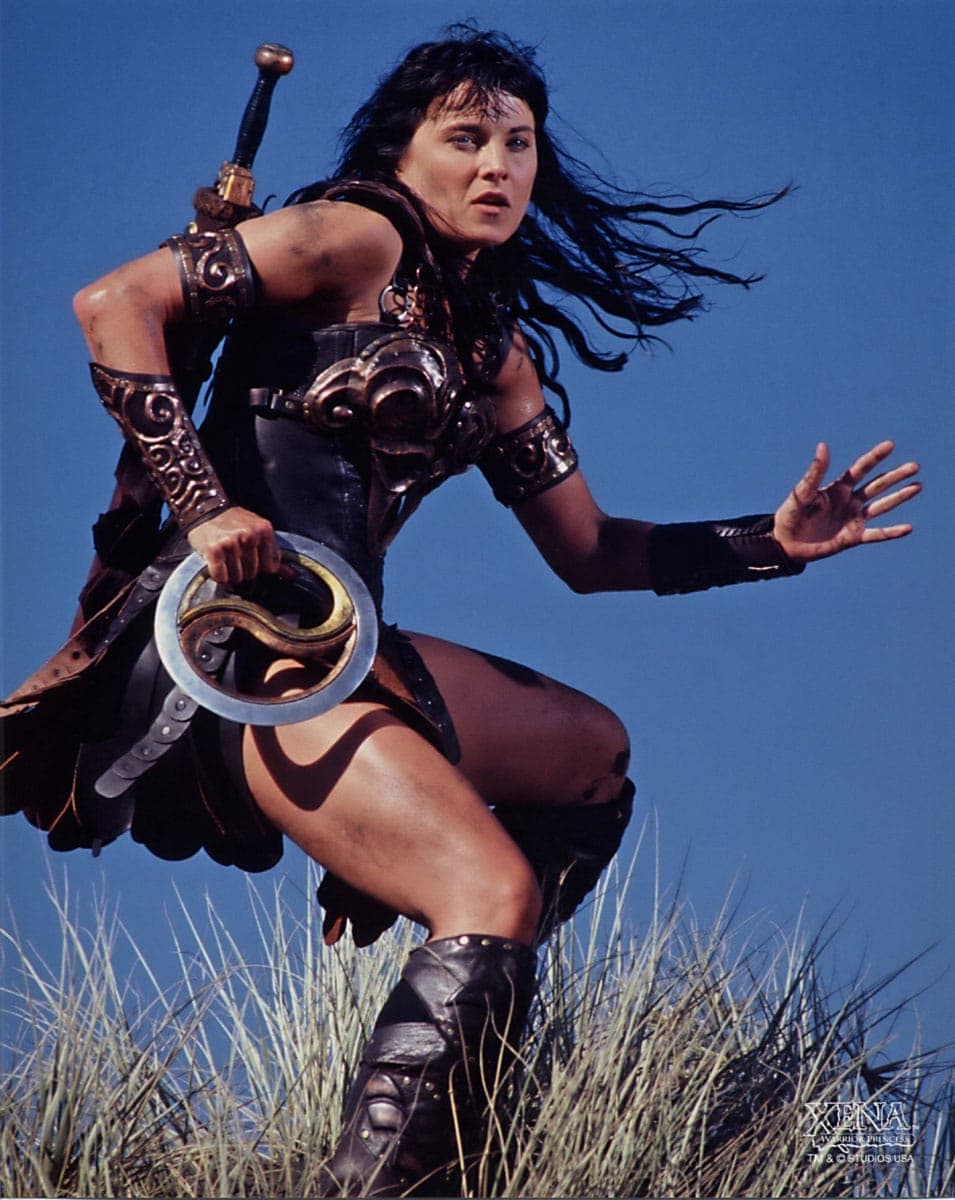 A promotional still of a woman in a leather warrior costume, holding a sword and a circular shield, running through tall grass against a blue sky.