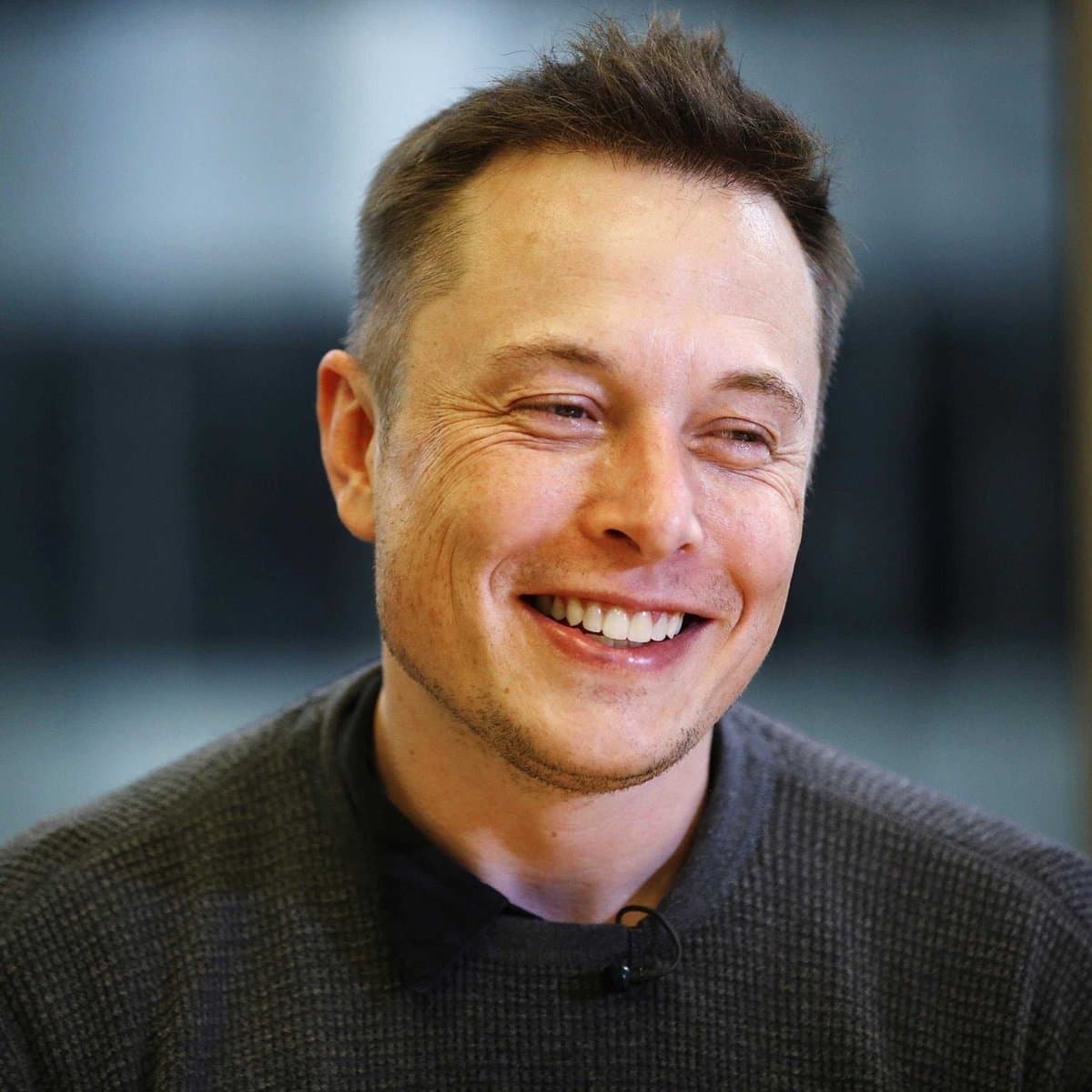 A close-up photograph of a smiling man with short brown hair and light stubble, wearing a dark gray sweater with a small lavalier microphone clipped to the collar. The background is blurred and out of focus.