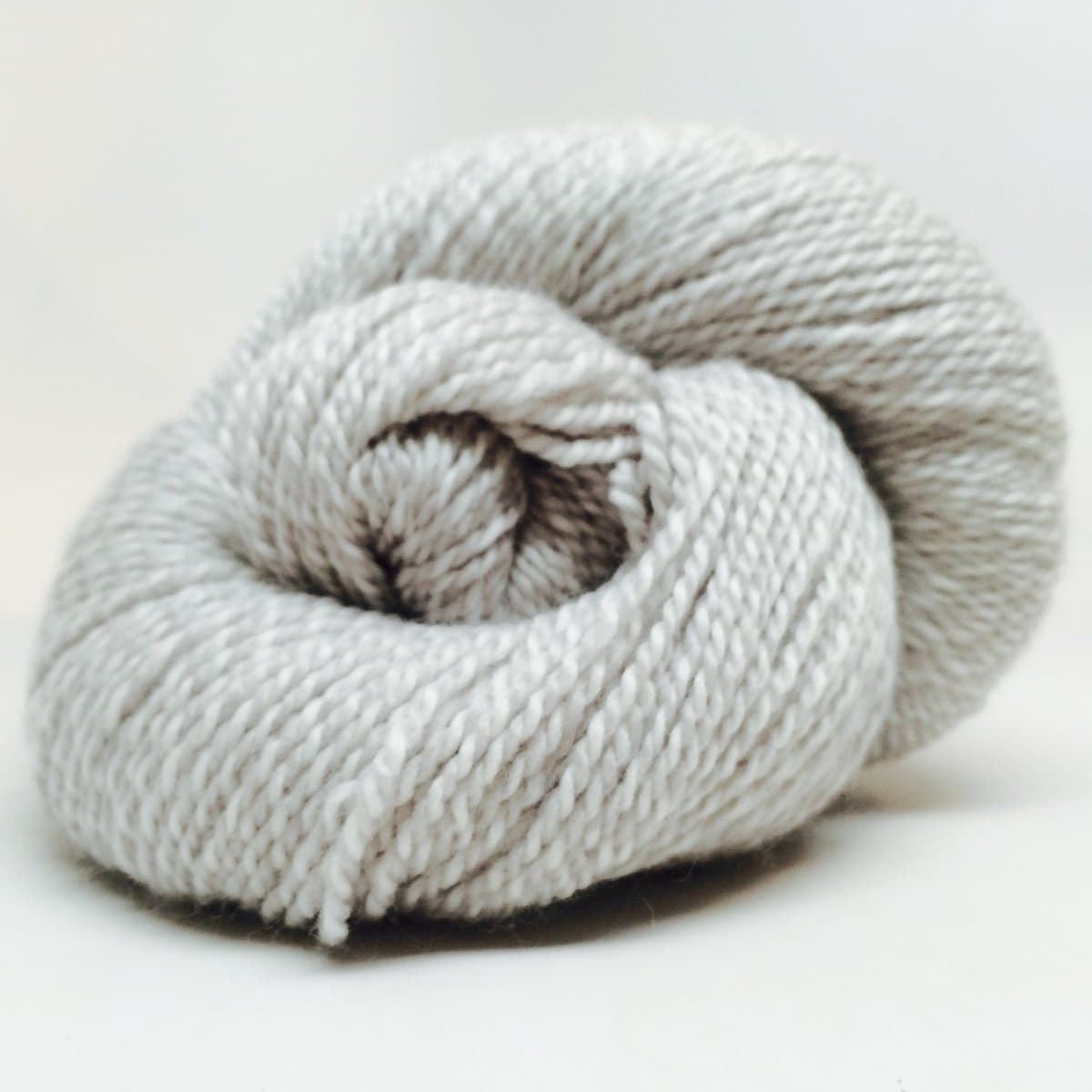 A close-up photograph of a skein of light gray or off-white yarn wound into a circular bundle, resting on a plain white surface. The yarn has a soft, slightly fuzzy texture typical of wool or a wool blend.