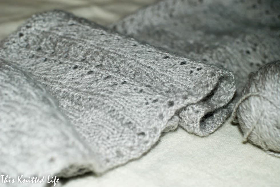 A close-up photograph of a soft, light gray knitted fabric with a textured pattern, draped over a white surface. A ball of matching gray yarn is visible on the right side of the frame.