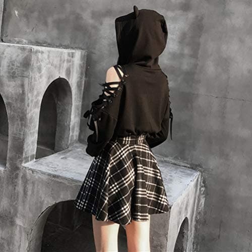 A person wearing a black hooded top with lace-up details and a black-and-white plaid skirt stands with their back to the camera in front of a gray concrete structure with arched openings.