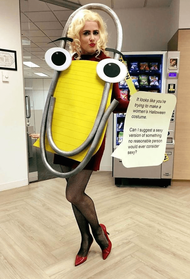 A woman in an office setting wears a Halloween costume made from a large yellow legal pad and a silver paperclip, with googly eyes attached to the pad. She is wearing a red top, fishnet tights, and red heels, standing in front of a snack vending machine.