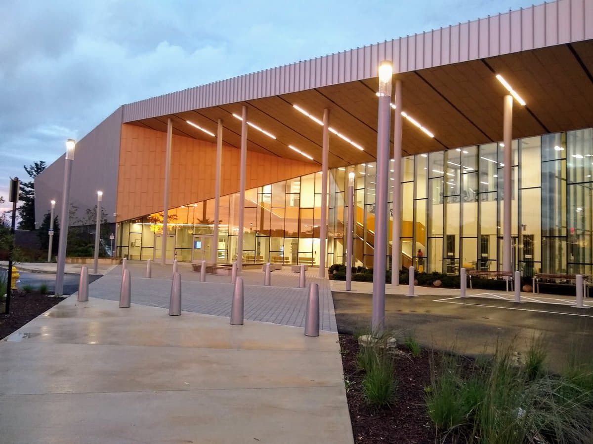An exterior view of a modern building with a large glass facade and a wooden overhang supported by tall, illuminated columns at dusk. The foreground features a paved walkway with metal bollards and a landscaped area with ornamental grasses.
