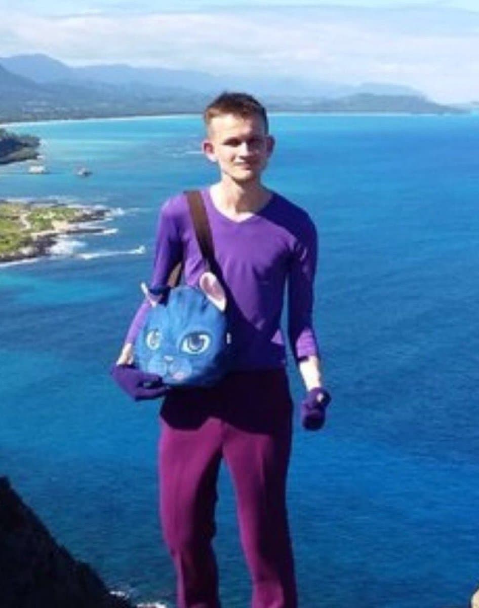 A person stands on a cliff overlooking a bright blue ocean and distant mountains, wearing a purple long-sleeved shirt and matching leggings. They are carrying a blue bag with a cartoon cat face design and wearing purple gloves.
