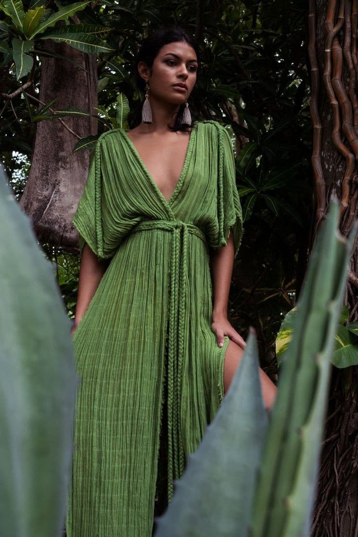 A woman stands outdoors among lush green foliage and trees, wearing a long, pleated green dress with a deep V-neckline and wide sleeves. She is accessorized with long, tassel earrings and looks directly at the camera.