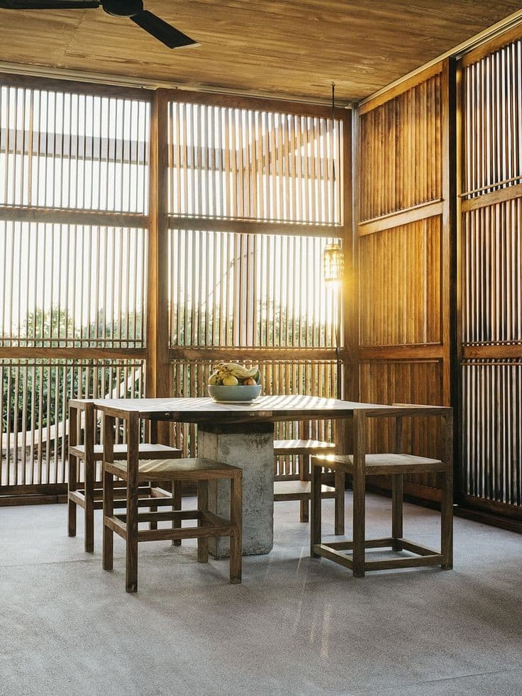 A photograph of a minimalist dining area featuring a wooden table and stools set on a concrete floor, surrounded by walls of vertical wooden slats that allow natural light to filter through. A bowl of fruit sits on the table, and a ceiling fan is visible overhead.