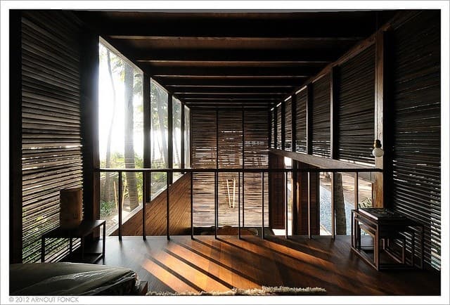 An interior architectural photograph of a modern, open-air wooden structure featuring slatted walls, a mezzanine level, and large glass windows looking out onto a tropical landscape. Sunlight streams through the windows, casting long shadows across the wooden floor.