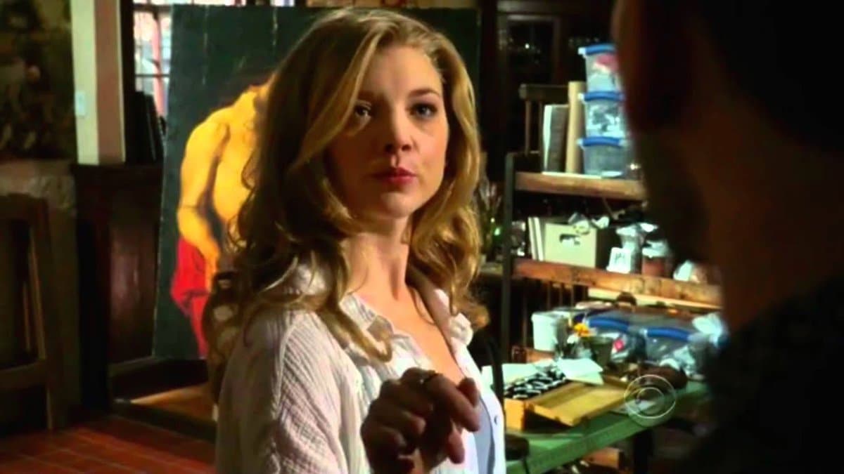 A still from a television show featuring a woman with wavy blonde hair wearing a white shirt, standing in a cluttered room with a large painting of a figure in the background.