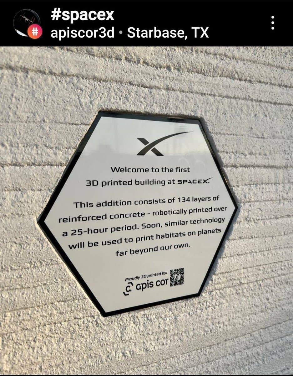 A photo of a hexagonal plaque mounted on a white, 3D-printed concrete wall. The plaque features the SpaceX logo and text announcing the structure as the first 3D-printed building at SpaceX, noting it consists of 134 layers of reinforced concrete printed over 25 hours.