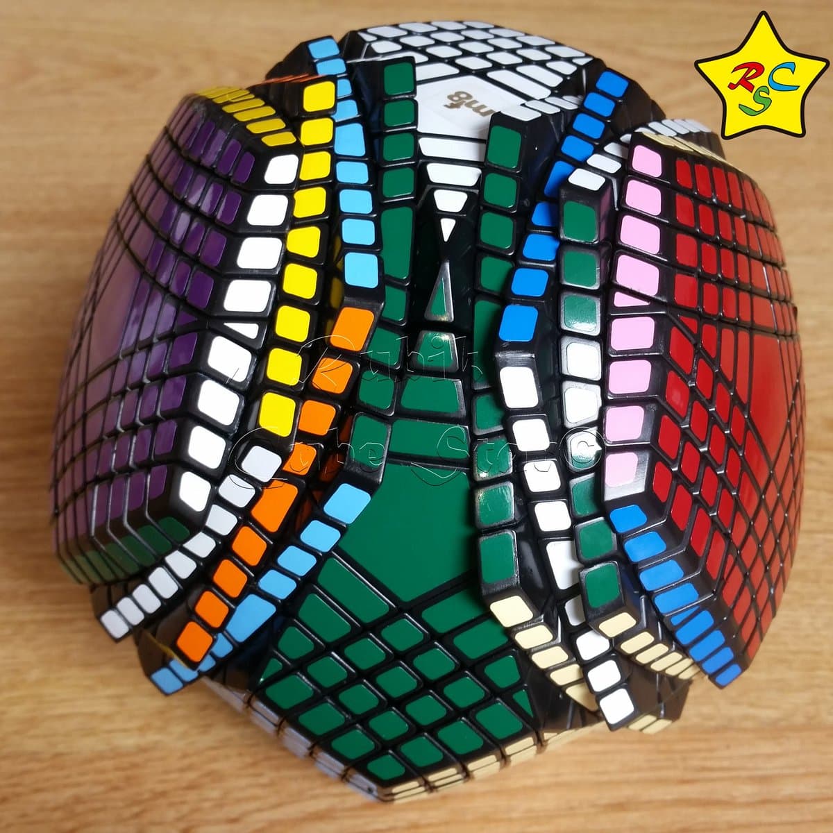 A photograph of a large, multi-layered twisty puzzle cube, likely a Pentamix, resting on a wooden surface. The puzzle features a complex grid of colored stickers in green, white, blue, red, orange, and purple, with a yellow star logo in the upper right corner.