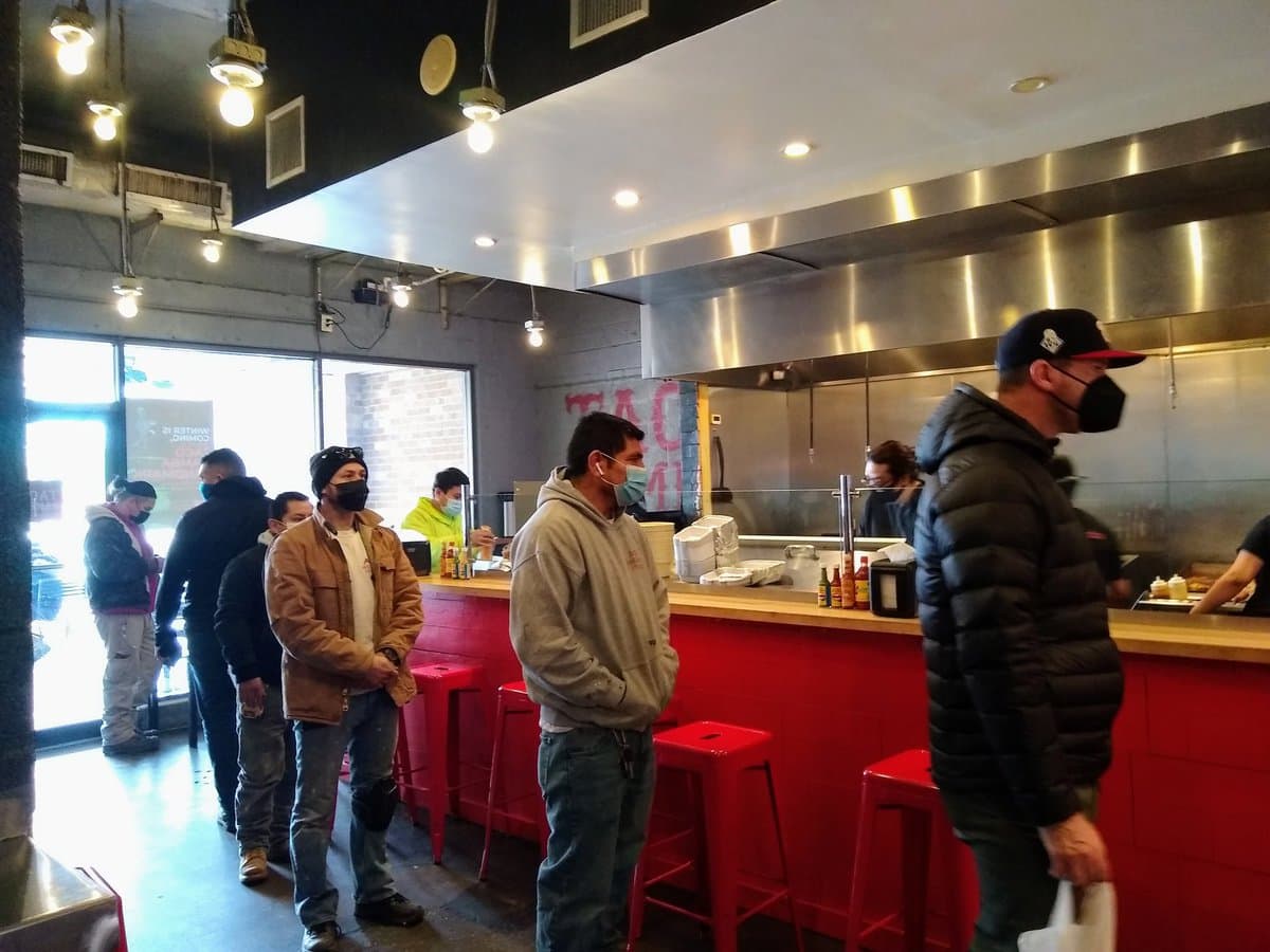 A photograph showing a line of people wearing face masks waiting at the counter of a restaurant with red stools and a stainless steel kitchen. A partially visible sign on the back wall reads "TACO BAMB...".