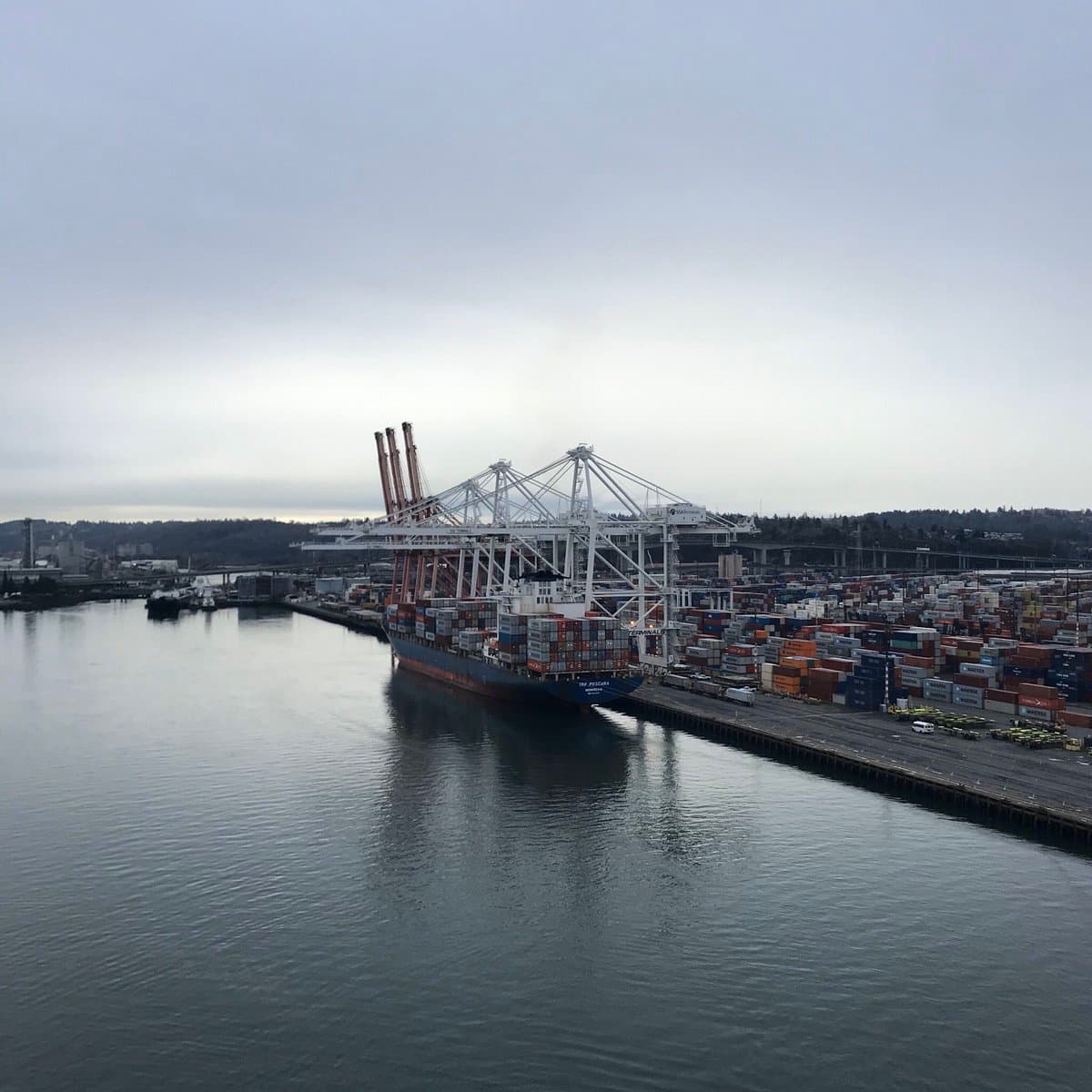 A wide-angle photograph of a container ship docked at a port terminal under an overcast sky. The vessel is positioned alongside a long pier equipped with large white gantry cranes, while the adjacent yard is filled with stacked shipping containers.