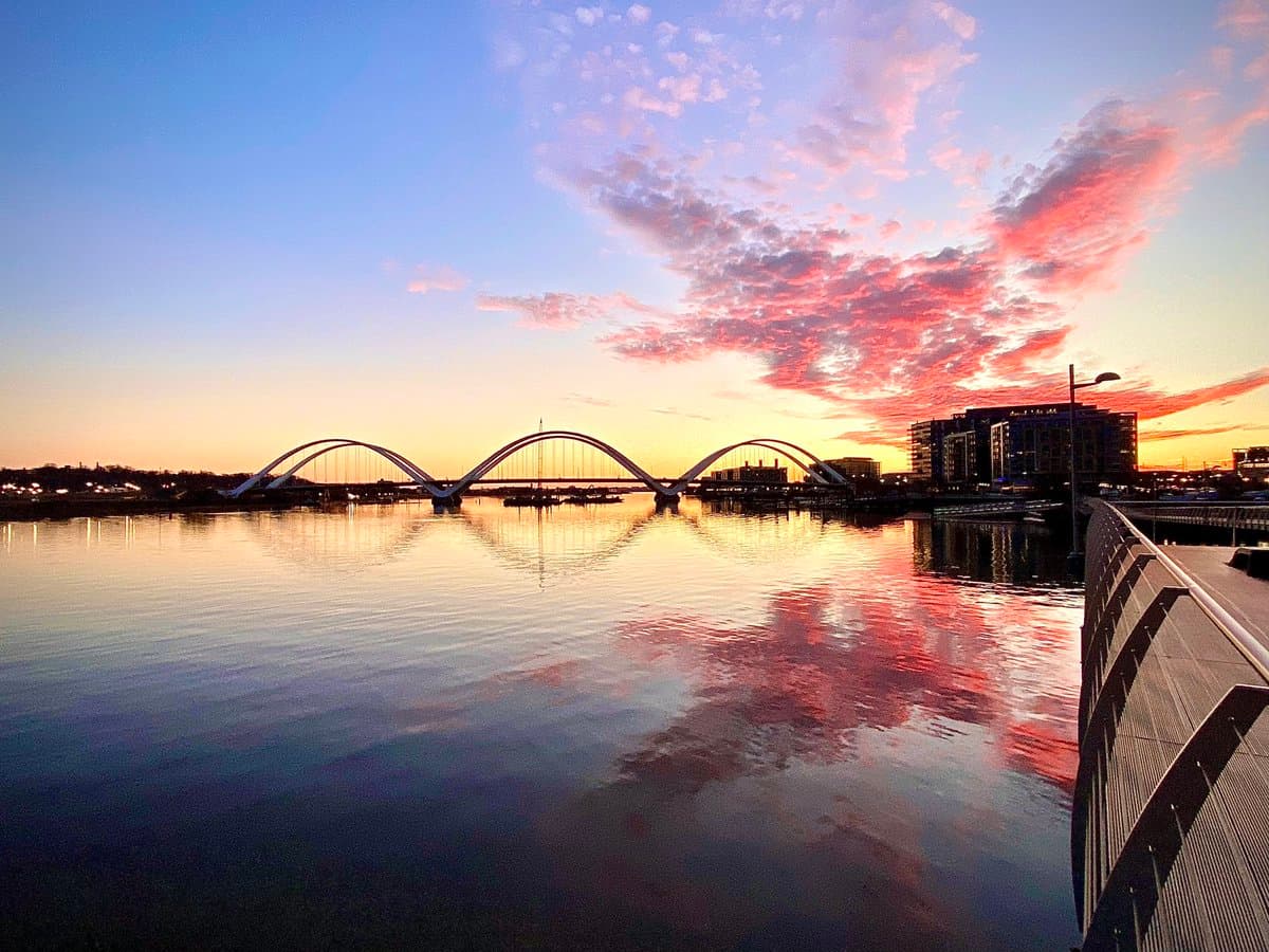 A scenic photograph of a city waterfront at sunset, featuring a distinctive white arch bridge spanning a calm river with a colorful sky reflected in the water.