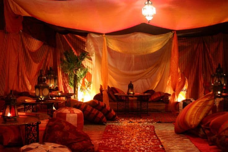 A dimly lit, exotic-style lounge or dining area decorated with flowing orange and red drapes, low seating with cushions, and lanterns. A patterned rug runs down the center of the floor, and a potted palm tree adds a touch of greenery to the warm, atmospheric setting.