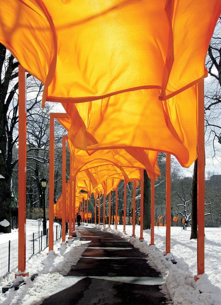 A long outdoor pathway is covered by a series of bright orange fabric canopies suspended from orange poles, creating a tunnel-like effect over a snow-covered walkway. Bare trees and a snowy landscape surround the installation, with a few people visible in the distance walking along the path.