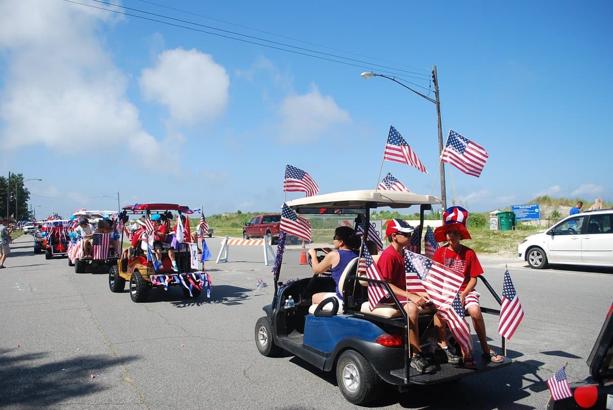 A line of decorated golf carts carrying people and numerous American flags travels down a paved road during a sunny day parade.