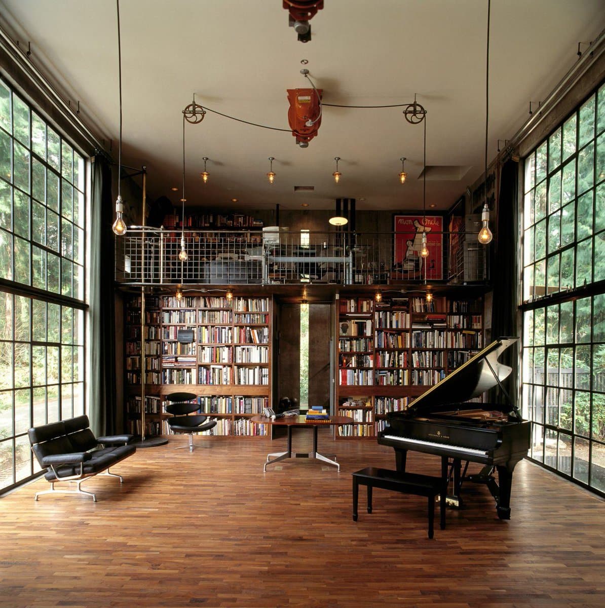 A wide interior photograph of a spacious living room featuring floor-to-ceiling windows, a mezzanine level, and walls lined with bookshelves. The room is furnished with a black grand piano, a black leather sofa, and a central table on a wooden floor.