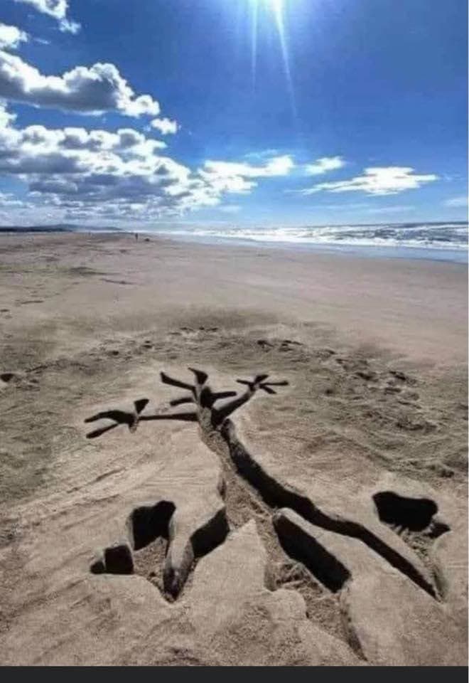 A photograph of a sandy beach under a bright, partly cloudy sky, featuring a large, dark, stick-figure-like drawing in the sand that resembles a person with outstretched arms and legs. The figure is positioned in the foreground, with the ocean and horizon visible in the background.