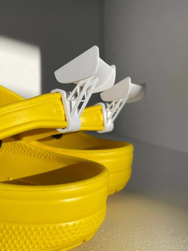 A close-up photograph of a pair of yellow Crocs sandals with white, 3D-printed aerodynamic wing attachments clipped onto the back of the straps.