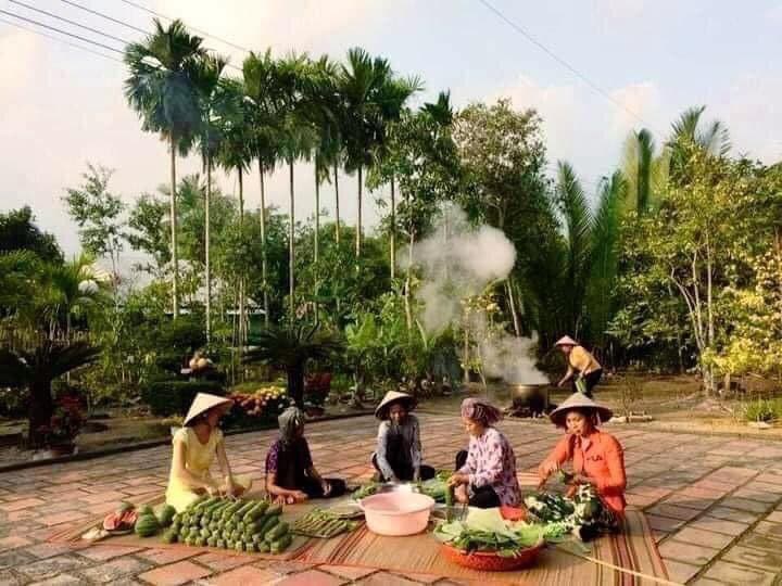 A group of people wearing traditional conical hats sit on mats on a paved outdoor area, preparing and selling fresh vegetables and produce. In the background, tall palm trees and lush greenery frame a scene where steam rises from a cooking pot, suggesting a local market or communal gathering in a tropical setting.