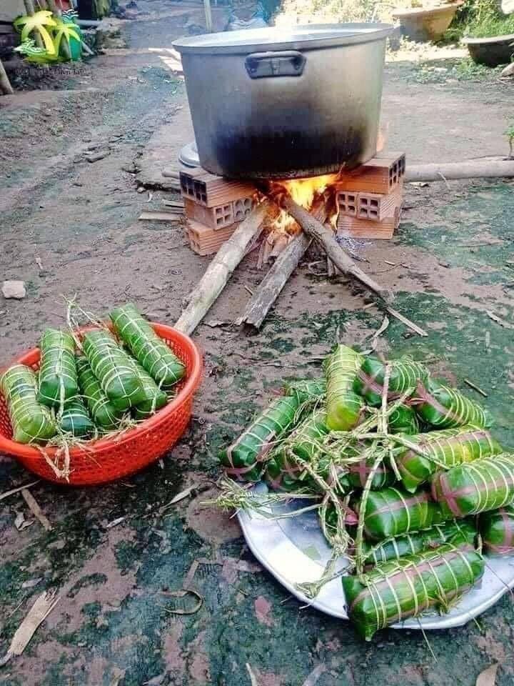 An outdoor scene showing a large metal pot boiling over a wood fire built on a brick base, with baskets and a plate of green, leaf-wrapped food parcels in the foreground.