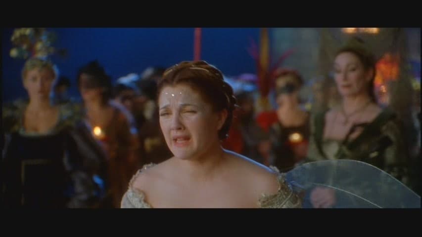 A still from a film or television show featuring a woman with reddish-brown hair in an updo, wearing an ornate, off-the-shoulder gown and a forehead jewel, with a distressed expression. She is surrounded by other people in a dimly lit, festive setting with blue and warm lighting.