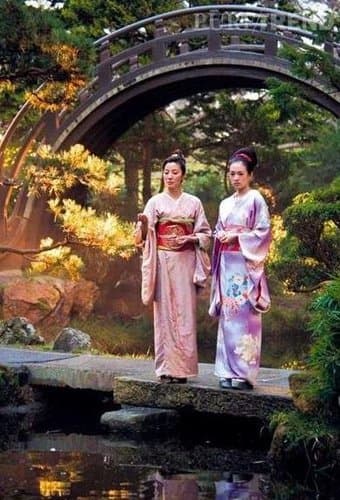 Two women wearing traditional Japanese kimonos stand on a stone path in a garden, with a wooden arched bridge and lush greenery in the background.