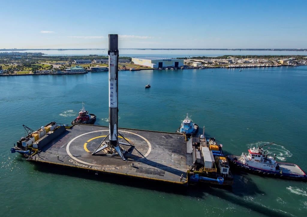 An aerial photograph shows a SpaceX Falcon 9 rocket standing vertically on a large floating barge in a body of water, surrounded by several tugboats. The barge has the text "Of Course I Still Love You" written on its deck, and the rocket is positioned on a circular landing pad.