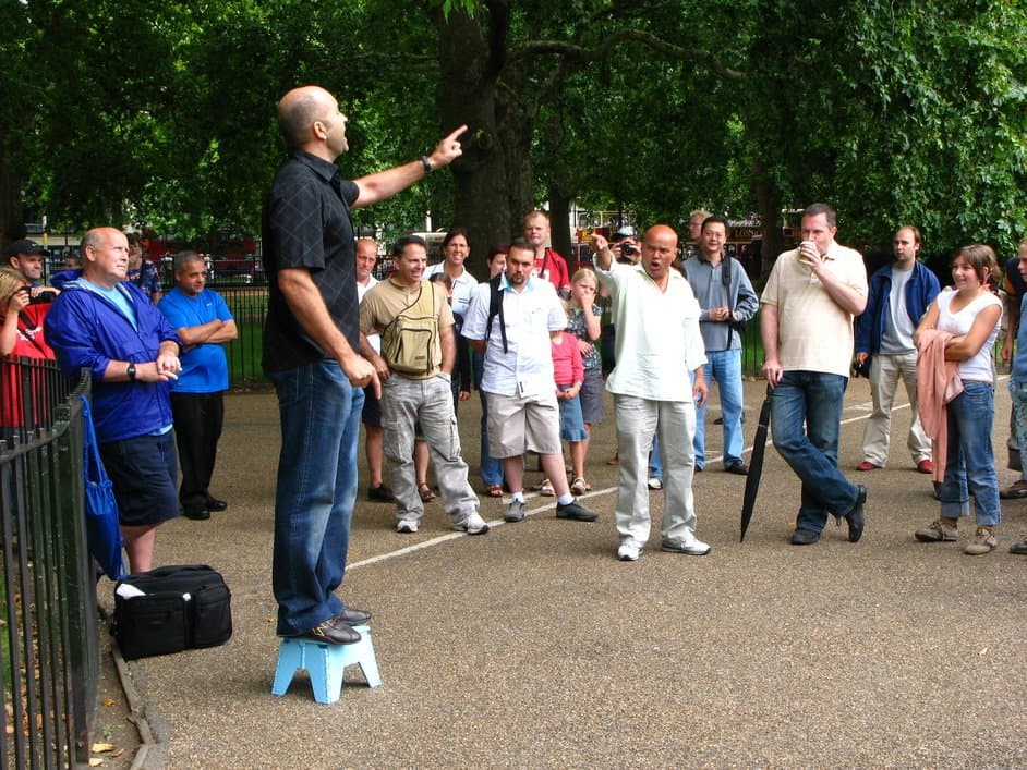 A man stands on a small blue stool in a park, pointing and speaking to a group of people who are gathered around him. The audience, consisting of men and women of various ages, listens attentively on a paved path surrounded by trees.