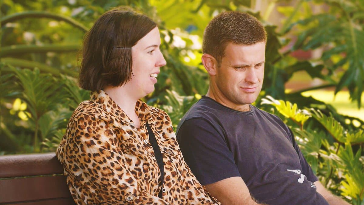 A woman in a leopard-print shirt and a man in a dark t-shirt sit on a bench outdoors, with the woman looking at the man who has a displeased expression. The background consists of green foliage.
