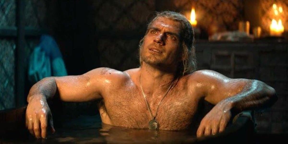 A shirtless man with long hair and a necklace is submerged in a dark, steaming bath, looking upward with a distressed expression. The scene is dimly lit by candles in the background, creating a moody and intense atmosphere.
