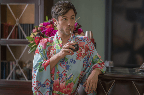 A man with short dark hair and a beard, wearing a colorful floral robe, sits indoors while smoking a hookah. He is positioned in front of a bookshelf and a vase of pink flowers.
