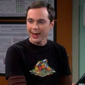 A man with short dark hair is smiling while wearing a black t-shirt featuring a graphic of a Rubik's Cube on a planet. He is standing indoors in front of a wall with a chart and text.