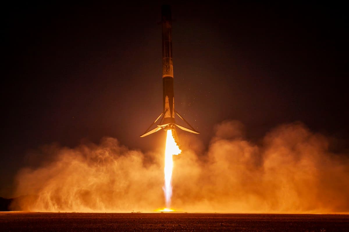 A Falcon 9 rocket booster is shown landing vertically on a flat, dark surface at night, with its landing legs deployed and engines firing to create a bright plume of fire and smoke. The scene is illuminated by the rocket's own exhaust against a dark sky.