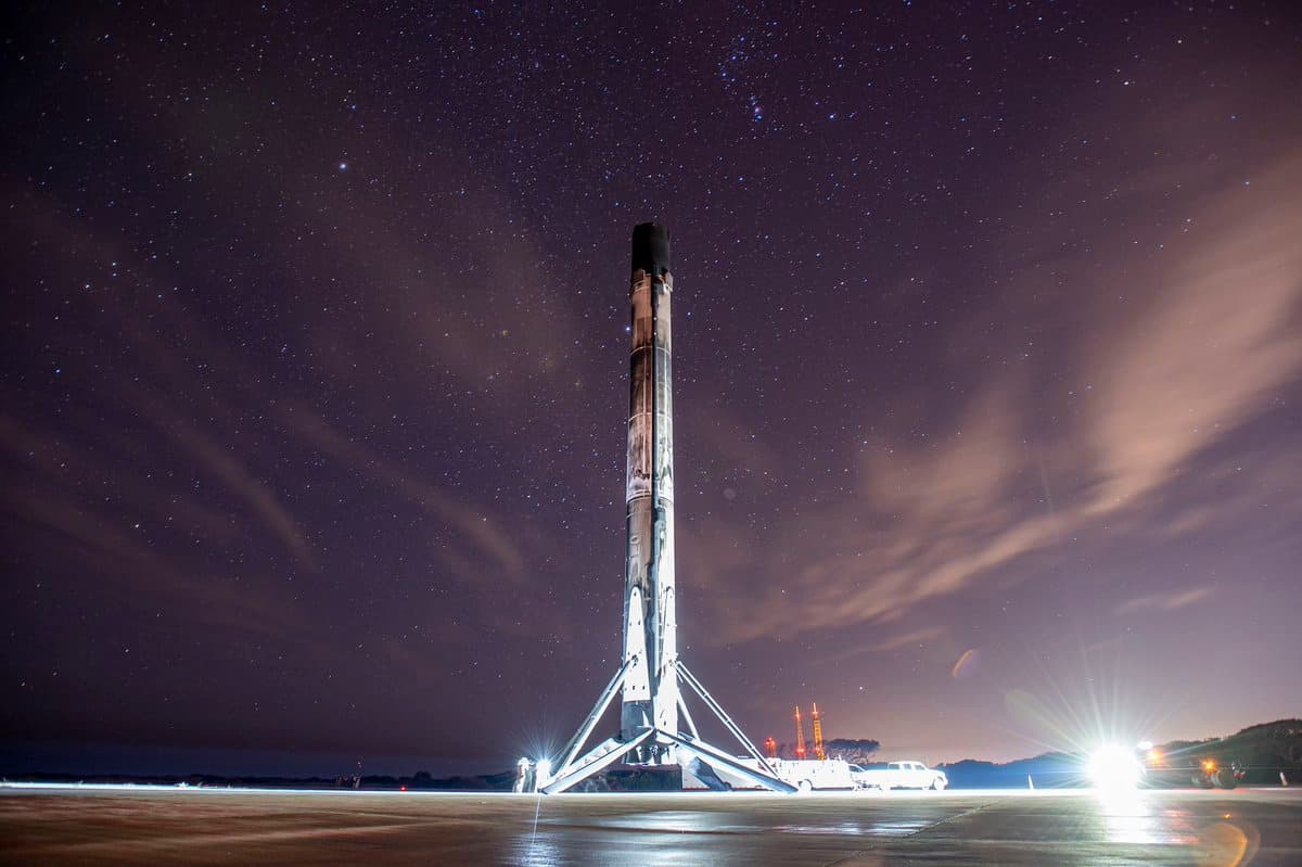 A Falcon 9 rocket booster stands vertically on its landing legs on a flat landing pad at night, illuminated by bright ground lights against a starry sky. The scene captures the moment after a successful landing, with support vehicles visible in the background.