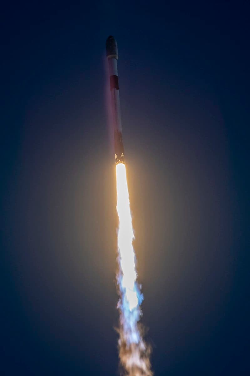 A Falcon 9 rocket ascends vertically against a dark sky, emitting a bright plume of fire and smoke from its base. The rocket's white body and dark interstage section are clearly visible as it climbs.