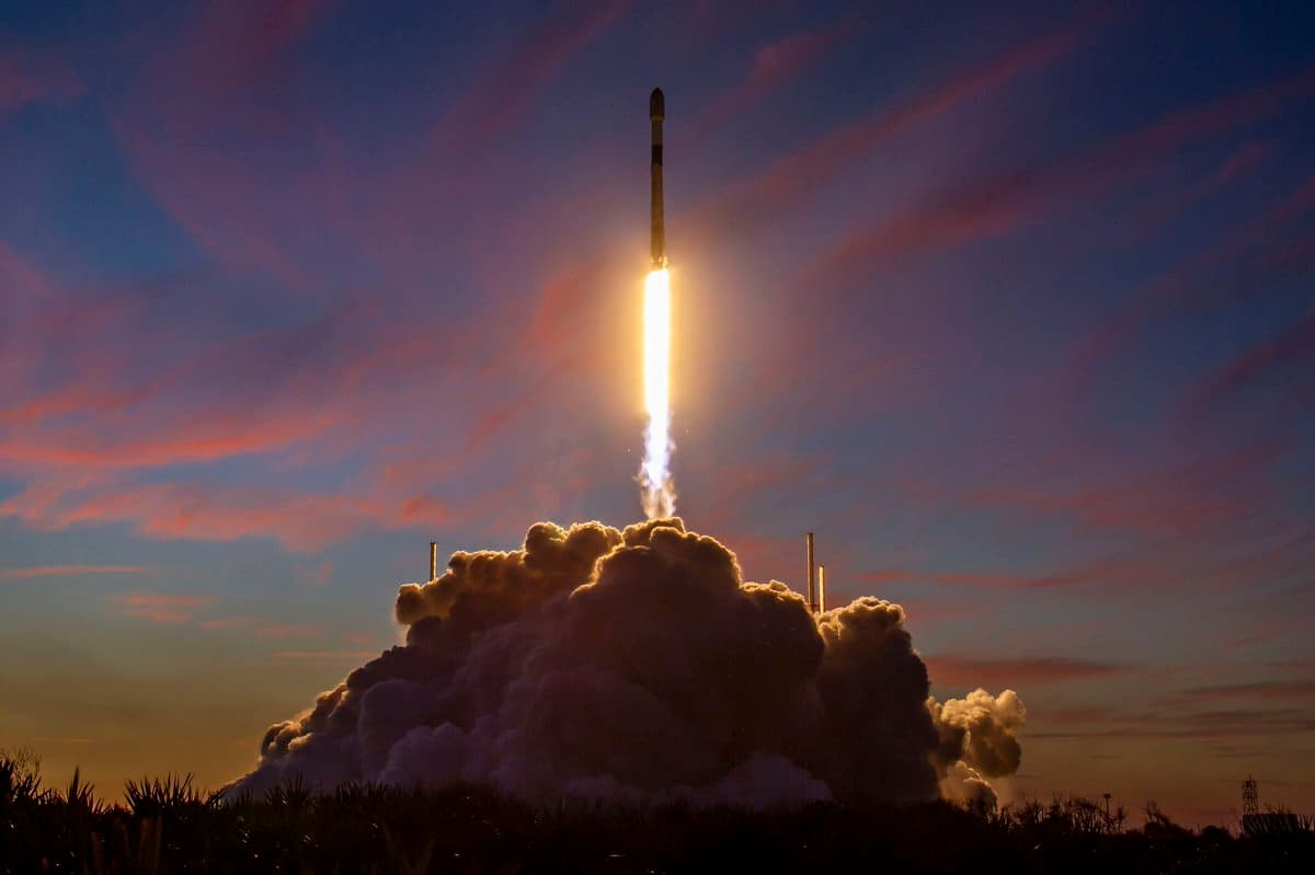 A rocket launches vertically into a twilight sky, emitting a bright plume of fire and smoke from its base. The scene is set against a backdrop of colorful clouds and a darkening horizon, with a large cloud of exhaust billowing at the launch site.