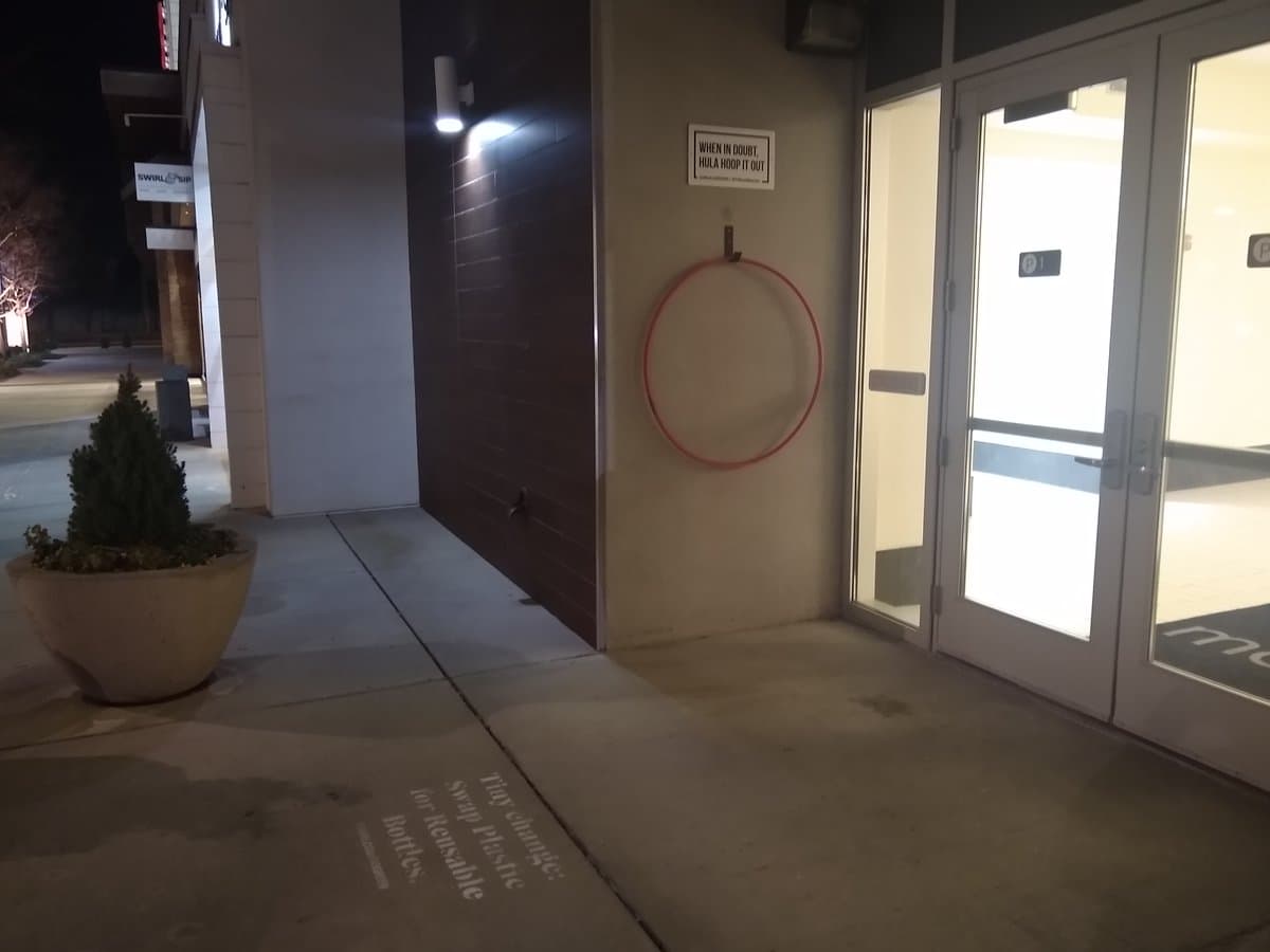 A nighttime photograph of a building entrance showing a red hula hoop hanging on a wall hook next to a sign. The scene includes a potted plant on the left and glass double doors on the right.