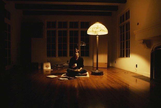 A person sits cross-legged on the floor of a dimly lit room, illuminated by a large, ornate floor lamp. The individual is surrounded by scattered papers and appears to be reading or writing. The room features high ceilings, large windows, and wooden flooring.
