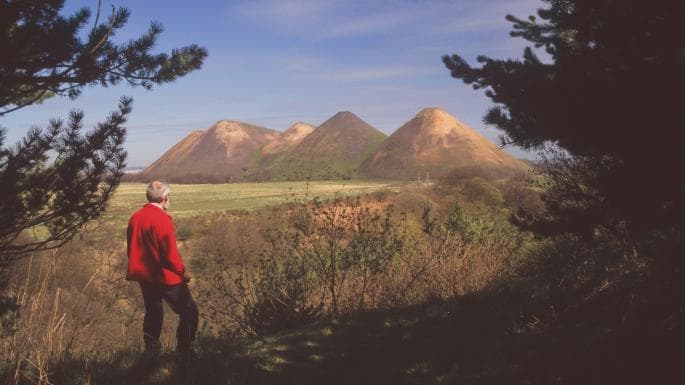 A landscape photograph showing a person in a red jacket standing in a field, looking out at a row of four large, conical spoil heaps in the distance under a blue sky.