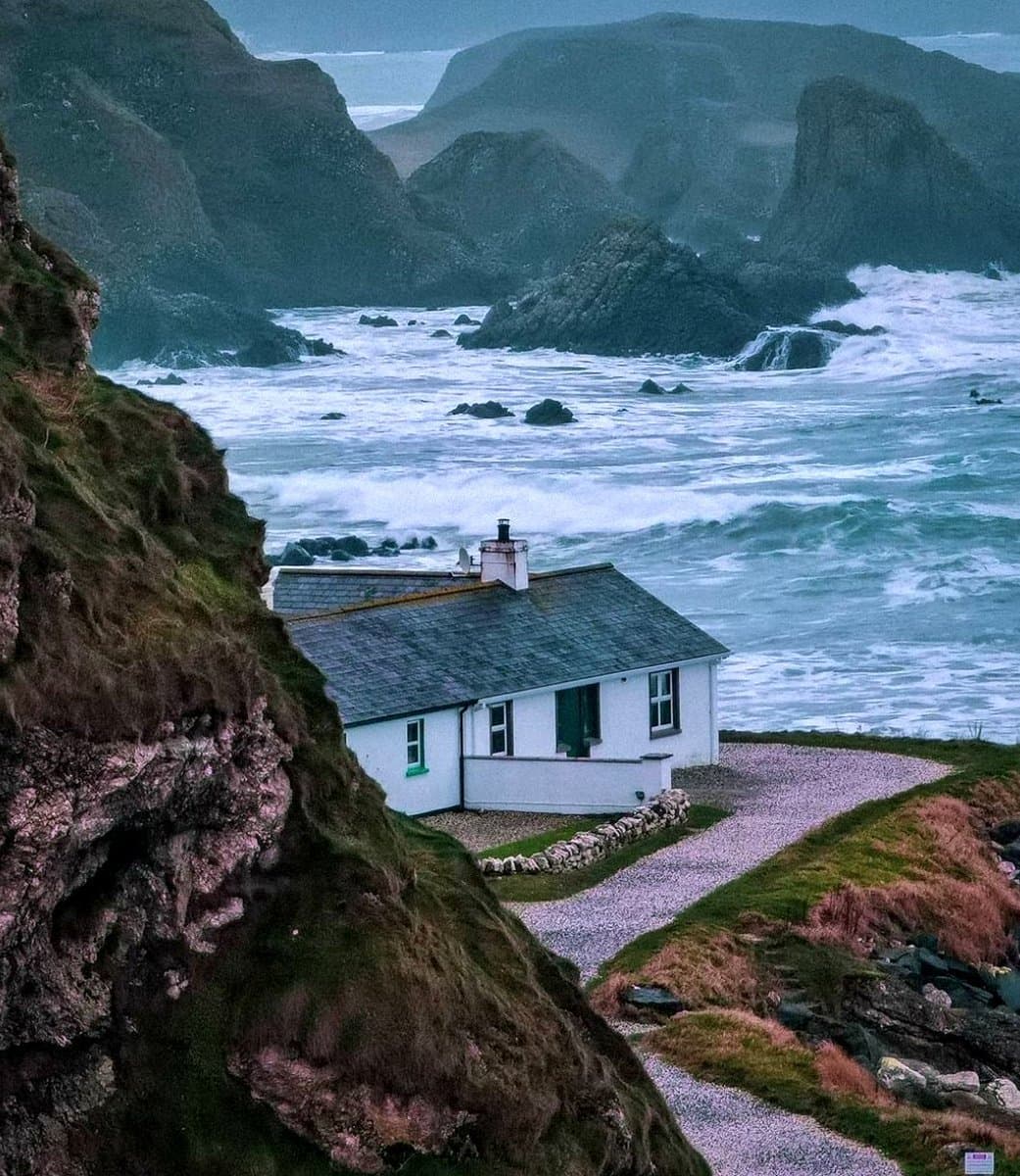 A white cottage with a slate roof sits on a grassy cliffside overlooking a turbulent, rocky ocean. The scene is framed by a steep, moss-covered cliff face in the foreground and rugged hills in the background under an overcast sky.