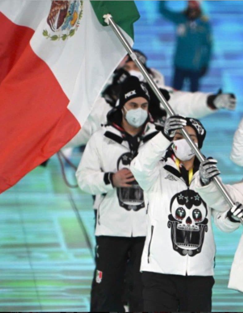 A photograph of two athletes from the Mexican Olympic team marching during a ceremony, wearing white jackets with a black sugar skull design and face masks while carrying the Mexican flag.