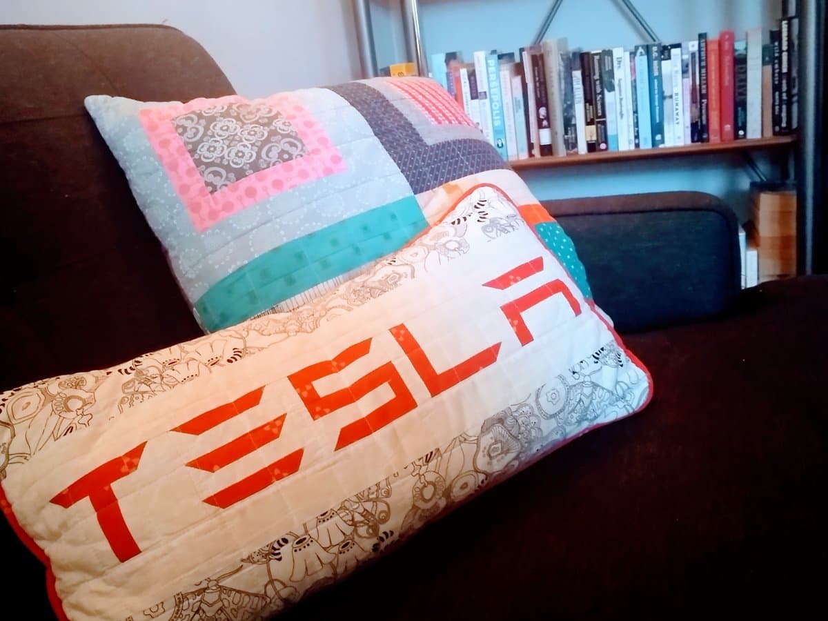 A photograph of two handmade, quilted pillows resting on a dark brown couch. The front pillow is white with a red 'TESLA' logo and a pattern of small robots, while the pillow behind it features a patchwork of pink, teal, and grey fabrics. A bookshelf is visible in the background.