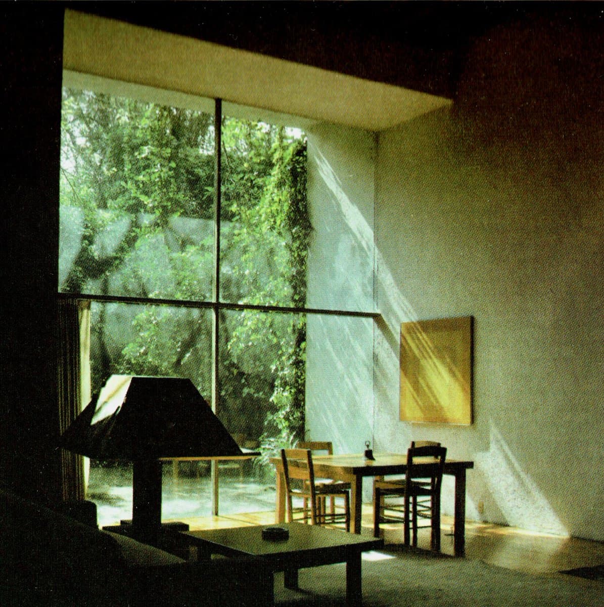 A photograph of a minimalist interior featuring a dining table and chairs positioned in front of a large window that looks out onto lush green foliage. Strong sunlight streams through the window, casting distinct shadows across the plain white wall where a square, yellow-toned piece of art is hung.
