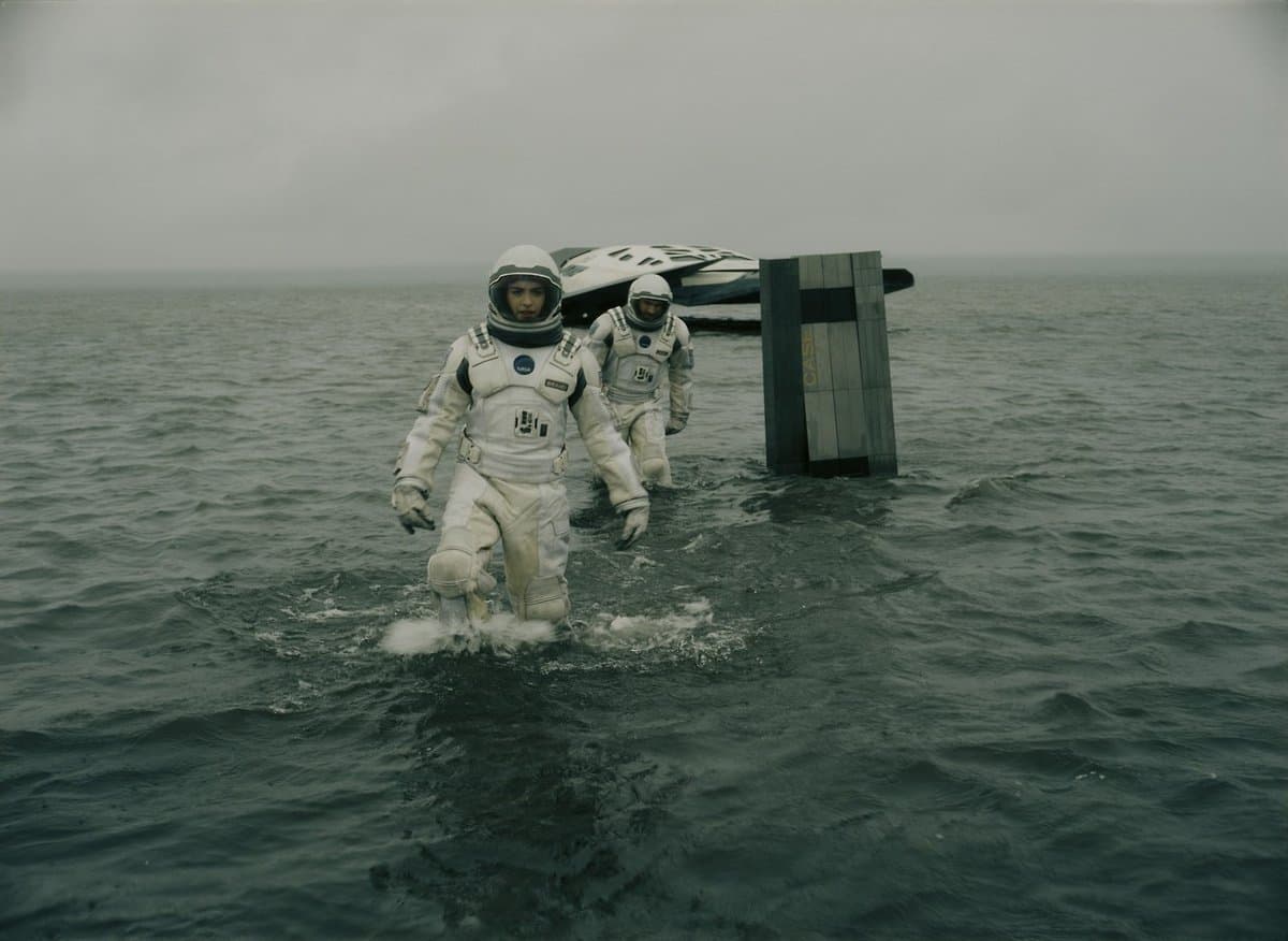 Two astronauts in white spacesuits wade through a calm, grey ocean toward the viewer, with a spacecraft and a vertical structure visible in the background under an overcast sky.