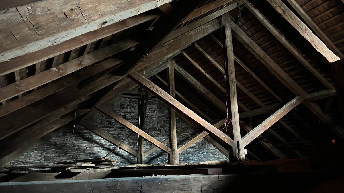 A low-angle photograph of an unfinished attic space featuring exposed wooden roof trusses and beams against a stone wall. The area is dimly lit, highlighting the texture of the aged wood and masonry.