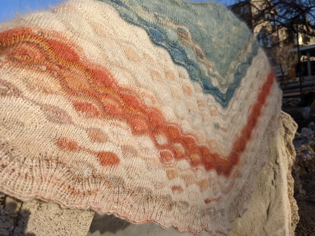 A close-up photograph of a hand-knitted shawl draped over a stone wall, featuring a textured pattern in cream, rust, and blue yarns. The background shows a residential street with houses and bare trees under a clear blue sky.
