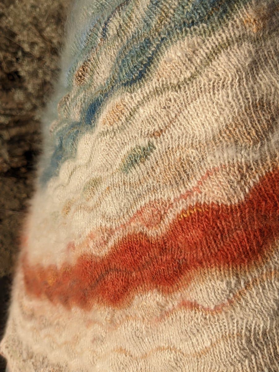 A close-up photograph of a hand-knitted textile featuring a textured stitch pattern. The fabric displays horizontal bands of color, including a prominent rust-orange stripe, a cream section, and a blue stripe near the top, resting on a fuzzy brown surface.